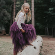 Young girl in a purple dress with a white lace top sitting on a log in a forest setting.
