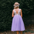 A young girl viewed from behind wearing a lavender tulle dress with a lace open-back bodice, standing outdoors in front of a dark green hedge.