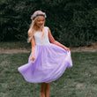 A young girl with a floral crown twirling in an outdoor grassy area, wearing a sleeveless white lace bodice and a voluminous lavender purple tulle skirt.