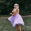 A young girl with a floral crown is photographed from behind outdoors on a grassy lawn, wearing a sleeveless lavender tulle dress with a lace bodice and a full flowing skirt.