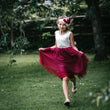 Young girl in a white top and red skirt running outdoors with greenery in the background