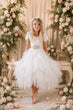 A young girl stands barefoot in a white lace and tulle flower girl dress, surrounded by a floral arch and candles.