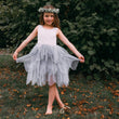 Young girl in a white top and gray tulle skirt standing outdoors with greenery in the background.