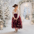 A young girl stands with her back to the camera in a snowy outdoor holiday setting, wearing a festive midi dress with a cream lace top and a deep burgundy tulle skirt decorated with gold floral embroidery.