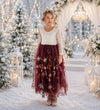 A young girl stands in a snowy outdoor ceremony setting wearing a long-sleeve white lace top and a burgundy tulle skirt with floral embroidery. The background features decorated Christmas trees, glowing string lights, and white lanterns.