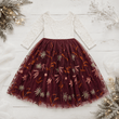A toddler girl's holiday dress featuring a white lace long-sleeve bodice and a full burgundy tulle skirt decorated with embroidered autumn leaves and pinecones, laid flat on a white wood background with festive holiday decorations.