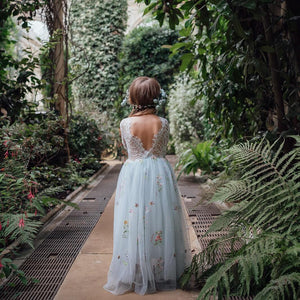 girl in a light blue embroidered dress walking through a lush green garden.