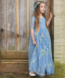 Young girl in a blue floral dress standing against a wooden wall.