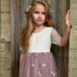 Young girl in a white lace top and purple floral dress standing against a stone wall.