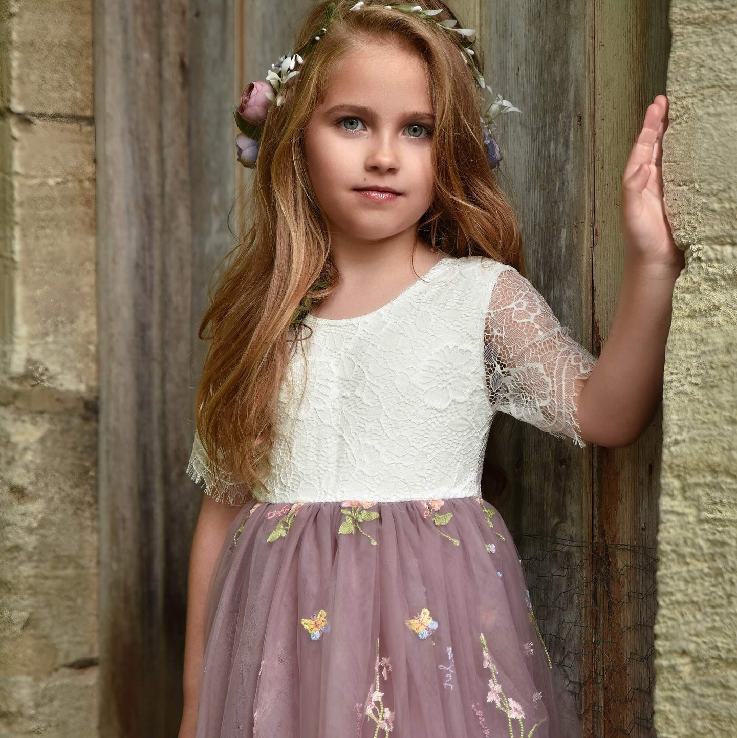 Young girl in a white lace top and purple floral dress standing against a stone wall.