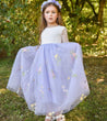 Young girl in a white top and light purple skirt with floral embroidery standing outdoors.