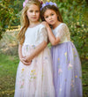 Two young girls in floral dresses standing outdoors with greenery in the background