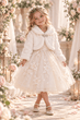 A young girl stands in a floral archway wearing a cream tulle dress with floral embroidery, a white faux-fur collar jacket, and a matching flower crown.