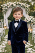 Young boy in a navy velvet suit standing in front of floral decorations at an outdoor event.