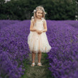 A young girl stands in the center of a lavender field wearing a sleeveless cream tulle dress with floral embroidery, a matching flower crown, and white sandals.