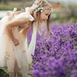 A young girl in a sheer cream tulle dress with floral prints and a matching flower crown leans over a field of purple lavender.