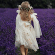 A young girl seen from behind wearing a white tulle dress with floral prints and a matching flower crown, standing in a field of purple lavender.