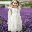 A young girl stands in a field of purple lavender wearing a white sleeveless dress with floral embroidery and a matching white headpiece.