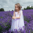 A young girl stands in a lavender field wearing a pale lavender tulle dress with a sheer matching veil and a floral crown, holding a bouquet of lavender stems.