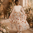 A young girl viewed from behind wearing a long-sleeve white dress with a brown and orange floral print skirt, standing in a rustic indoor setting decorated with flowers, candles, and warm string lights.