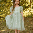 Young girl in a light green dress with floral embroidery standing in a forest setting.