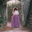 Girlin a white top and purple skirt standing in a stone archway.