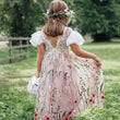Child in a floral dress standing in a grassy field with a wooden fence and trees in the background.