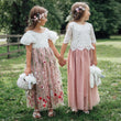Two young girls in floral dresses holding hands in a grassy outdoor setting.