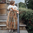 Young girl in a vintage dress standing on stone steps outdoors with a house and trees in the background.