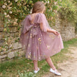 A young girl viewed from behind wearing a dusty purple tulle dress with floral embroidery, puff sleeves, and a large bow at the back, standing outdoors in front of a stone wall with climbing flowers.