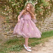 Young girl in a enchanted pink floral dress standing in front of a stone wall with flowers.