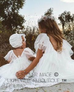 Two young girls in white dresses sitting outdoors with trees in the background.