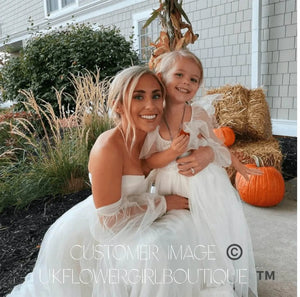 Woman and young girl in white dresses sitting outdoors with pumpkins and hay bales.