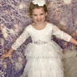 A young girl smiling in a white lace and tulle flower girl dress with a jeweled waistband, standing against a snowy forest backdrop. She has a white floral headband.