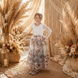 A young girl stands in a styled boho room wearing a white long-sleeve top and a long white skirt with a brown and gray botanical pattern. The background features a macrame curtain, pampas grass, and dried floral arrangements.