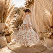 A young girl stands with her back to the camera wearing a white bohemian-style maxi dress with a floral print and lace sleeves, set in a warm, decorated outdoor scene with pampas grass, macrame, and dried flowers.
