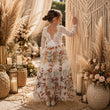 A young girl stands with her back to the camera in a bohemian-style decorated hall, wearing a white lace long-sleeve dress with a floral print skirt. The setting features a macrame wall hanging, pampas grass, candles, and woven baskets.