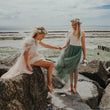 Two girls sitting on rocks by the ocean, holding hands wearing dresses from UK Flower Girl Boutique