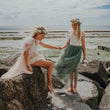 Two girls sitting on rocks by the ocean, holding hands.