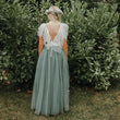 A woman stands with her back to the camera wearing a flowing dusty sage green tulle dress with a white sheer back bodice and a white floral crown, set against a background of green garden bushes.