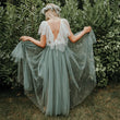 A woman stands outdoors in front of dense green foliage, viewed from behind, wearing a flowing tulle dress in a muted sage green color. The dress has a deep open back, short sheer sleeves, and a sheer waist tie.