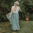 A woman stands barefoot on grass in a garden, photographed from behind, wearing a flowing sheer tulle dress in a muted sage green color with a white floral crown.