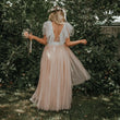 A woman stands outdoors in front of dense green foliage, photographed from behind, wearing a flowing blush pink tulle dress with sheer white flutter sleeves, a white waist tie, and a floral crown. She is holding a small bouquet.