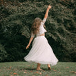 A woman wearing a white lace crop top and a flowing white tulle midi skirt, captured from behind as she twirls barefoot on a grassy lawn with a dense green hedge in the background.