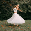 A young girl twirling outdoors in a white lace crop top and a voluminous white tulle skirt, set against a background of green trees and grass with scattered brown leaves.