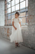 A young girl wearing a white tulle first communion dress with lace sleeves, standing in a rustic industrial room with brick walls and a large window.