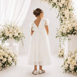 A young girl viewed from behind wearing a white tulle flower girl dress with lace puff sleeves and a bow on the back, standing under a floral archway.