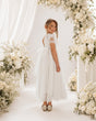 A young girl stands in a white floral arch wearing a white tulle flower girl dress with lace cap sleeves and metallic silver shoes, set against a bright white background with draped fabric and white flowers.