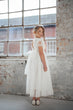 A young girl stands in a bright, industrial room with a large window and exposed brick wall, wearing a long white tulle dress with lace sleeves and white shoes.