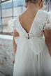 A child wearing a white lace and tulle dress, shown from the back. The dress features a deep V-back, floral lace applique, ruffled short sleeves, and a large bow at the back with a decorative bead detail.